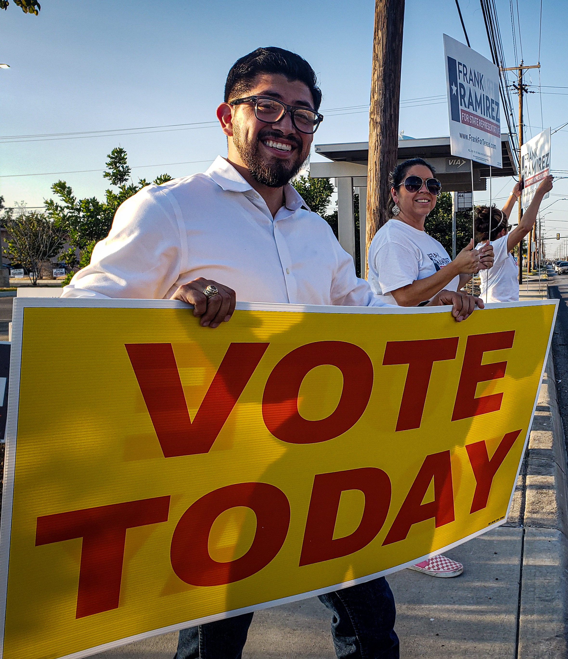 Frank Ramirez in a white shirt standing on the side of a busy street holding a yellow sign with red lettering that reads 'Vote Today'. A supporter in sunglasses stands a few feet to his left sporting a large smile and giving a thumbs up.